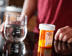 Primer plano de un frasco de pastillas y un vaso de agua con un hombre de fondo.